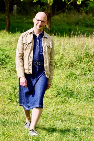 Mann in einem Jeanskleid steht lächelnd auf einer Wiese, umgeben von grüner Natur.