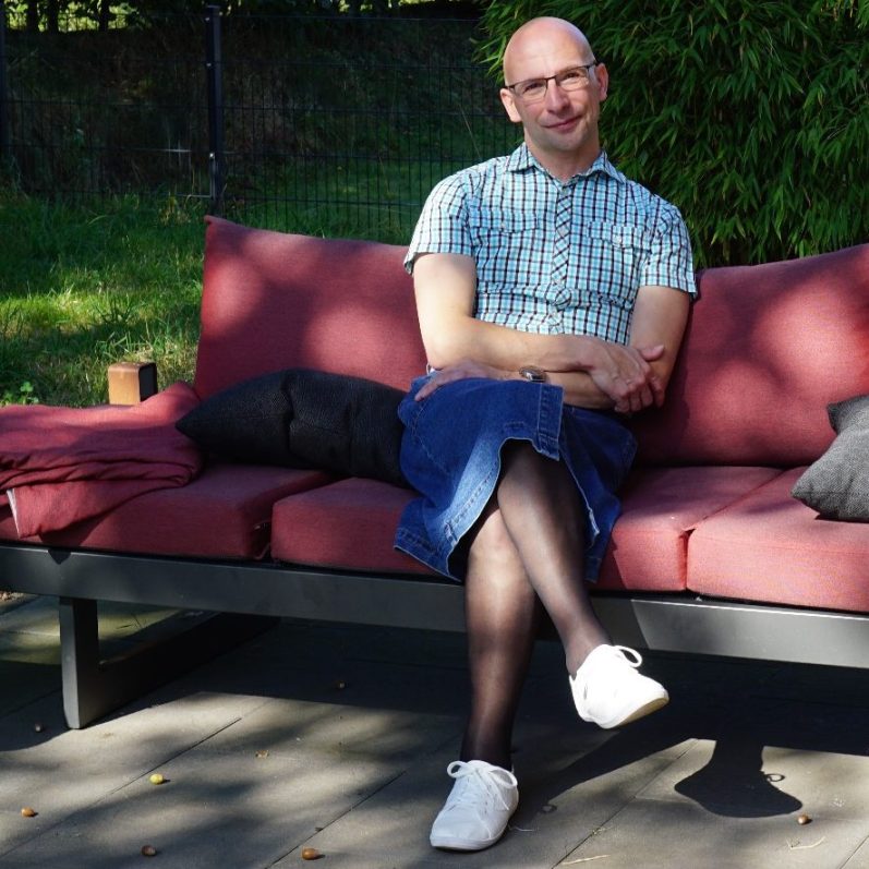 Mann in kariertem Hemd und Rock sitzt entspannt auf einer Couch im Freien.