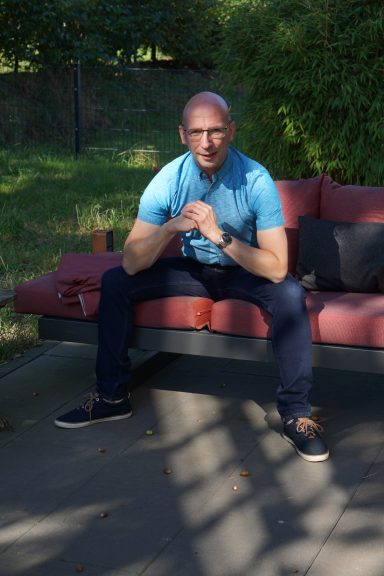 Mann sitzt auf einer Bank, trägt ein blaues Shirt und schaut direkt in die Kamera.