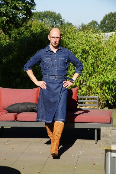 Mann in einem Jeanskleid und Stiefeln steht lässig auf einer Terrasse.