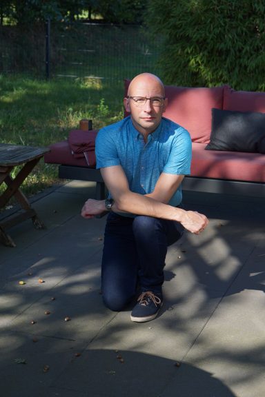 Mann in blauer Hemd und dunkler Hose kniet auf einer Terrasse.