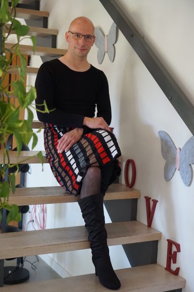 Person mit Brille, schwarzem Oberteil und gestreiftem Rock sitzt auf Treppe in einem modernen Raum.