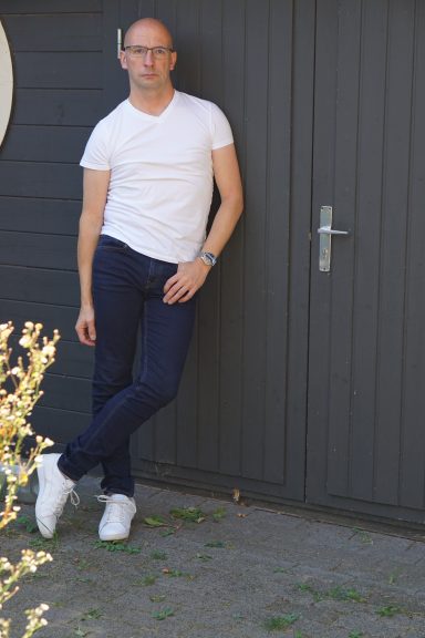 Mann mit Brille in weißem T-Shirt und Jeans lehnt an einer grauen Wand.